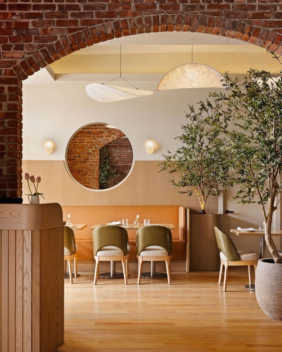 Clementina dining room with exposed brick arch, maple restaurant tables, brushed stainless steel table bases, natural wood chairs, olive green upholstery, cognac banquette seating, and indoor greenery.