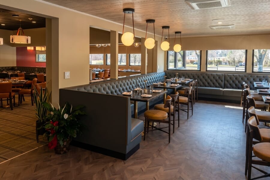 Window-side dining area with bar height tables, upholstered bar stools, antique black table tops, and warm wall lighting.