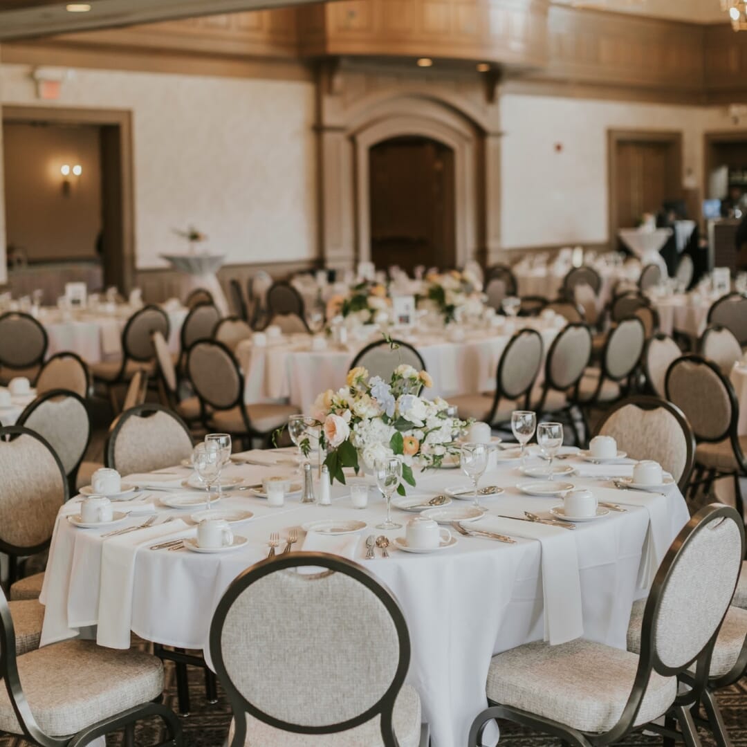 Rush Creek Golf Club MacMillan Ballroom featuring Eliseo teardrop-back banquet chairs around wedding reception tables, a large stone fireplace, vaulted ceiling, and panoramic windows.