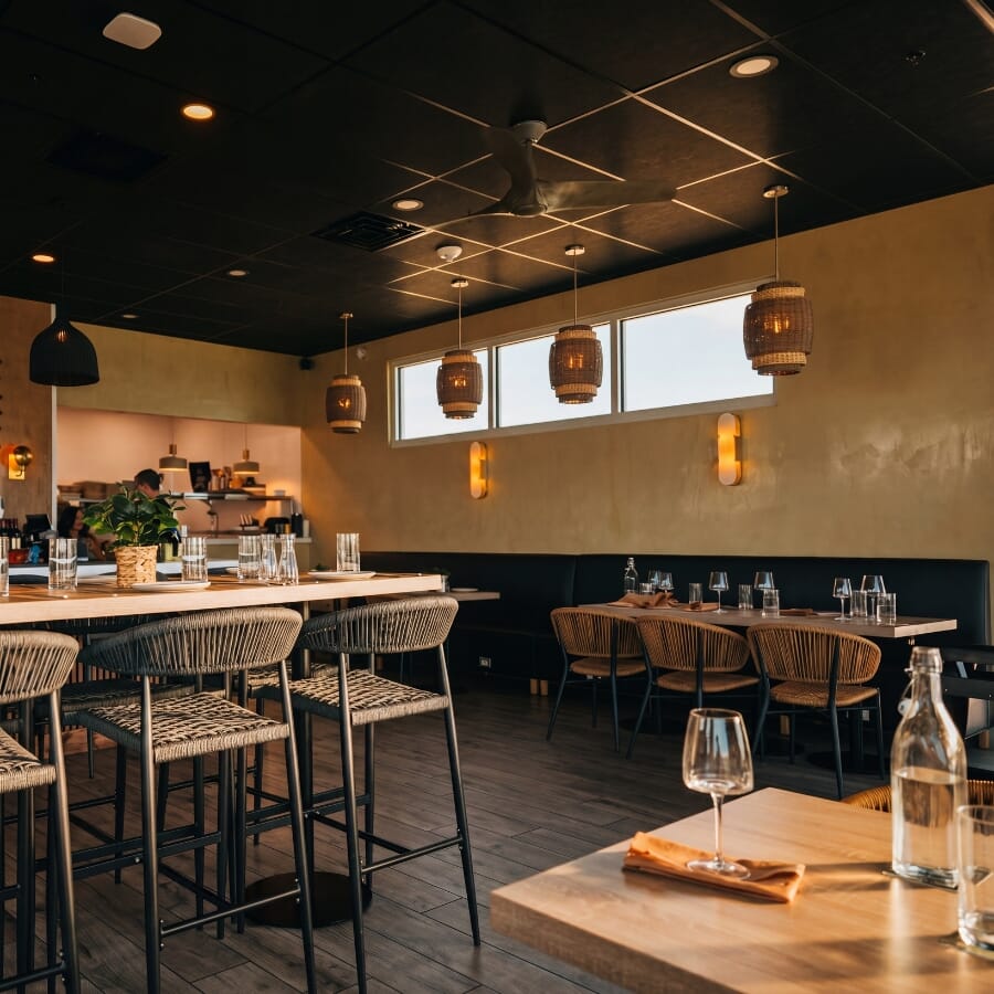 Coastal restaurant dining area with tan rope chairs, oak table tops, black round table bases, and warm neutral finishes.