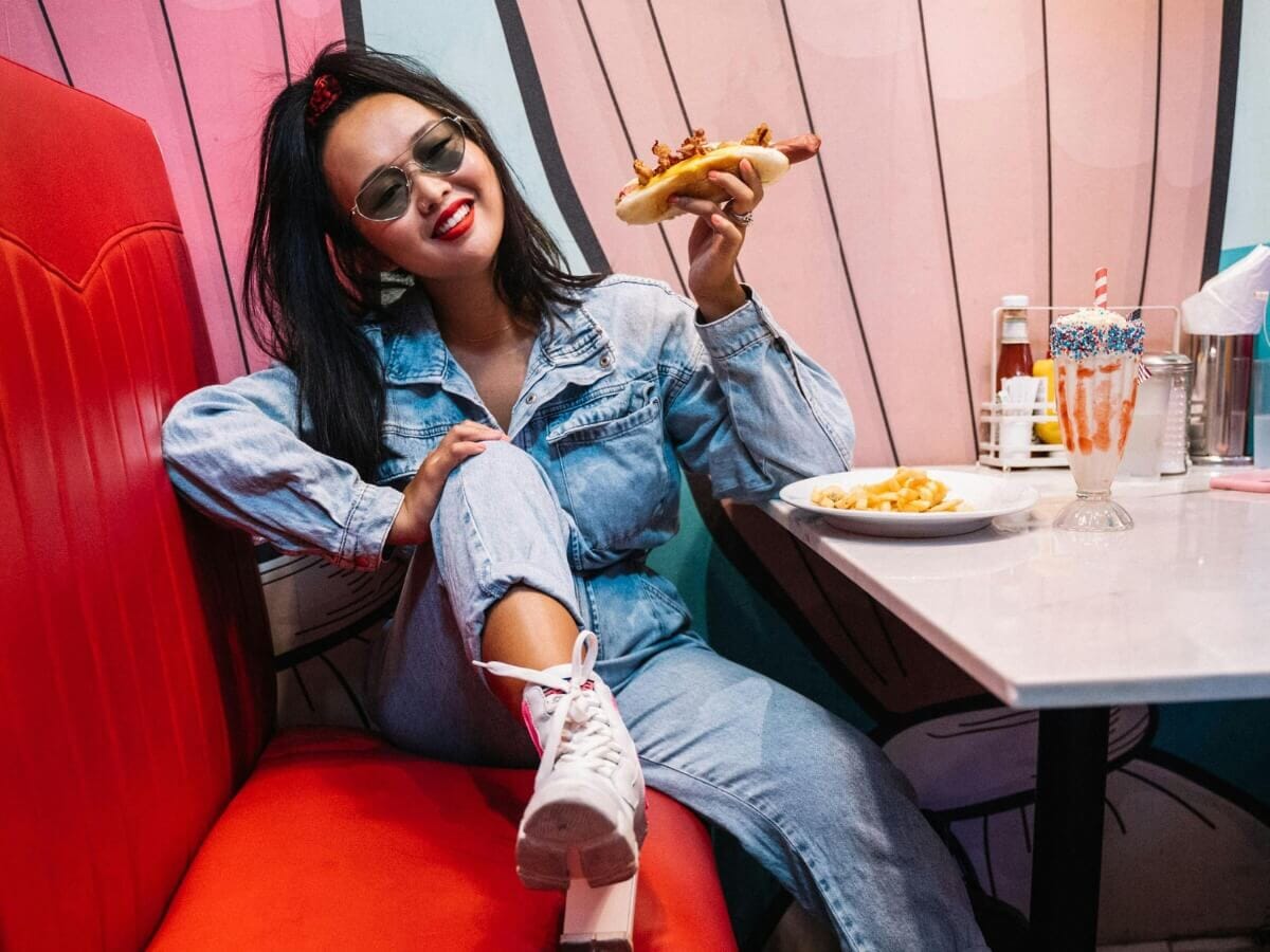 A young woman social influencer wearing denim and sunglasses enjoying a hot dog in a retro diner, illustrating modern restaurant social media marketing trends.