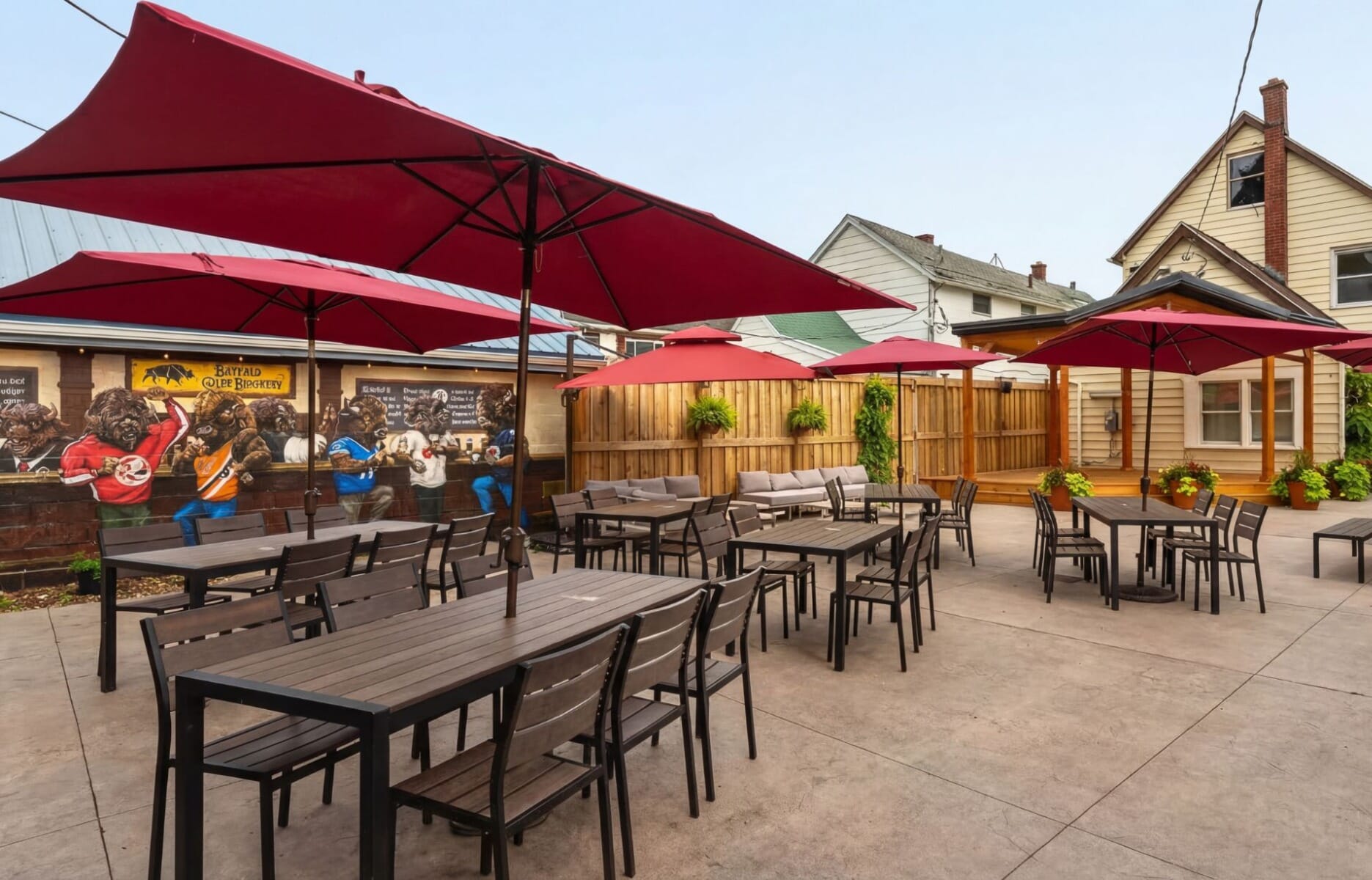 Outdoor patio at Buffalo Olde Brewery in Buffalo New York featuring dark wood tables, red market umbrellas, lounge seating, and a colorful Buffalo Bills sports mural on the exterior wall