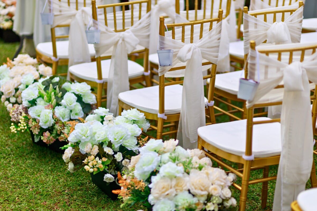 Gold Chiavari chairs with white cushions and draped fabric bows, decorated with floral arrangements for an outdoor wedding ceremony.