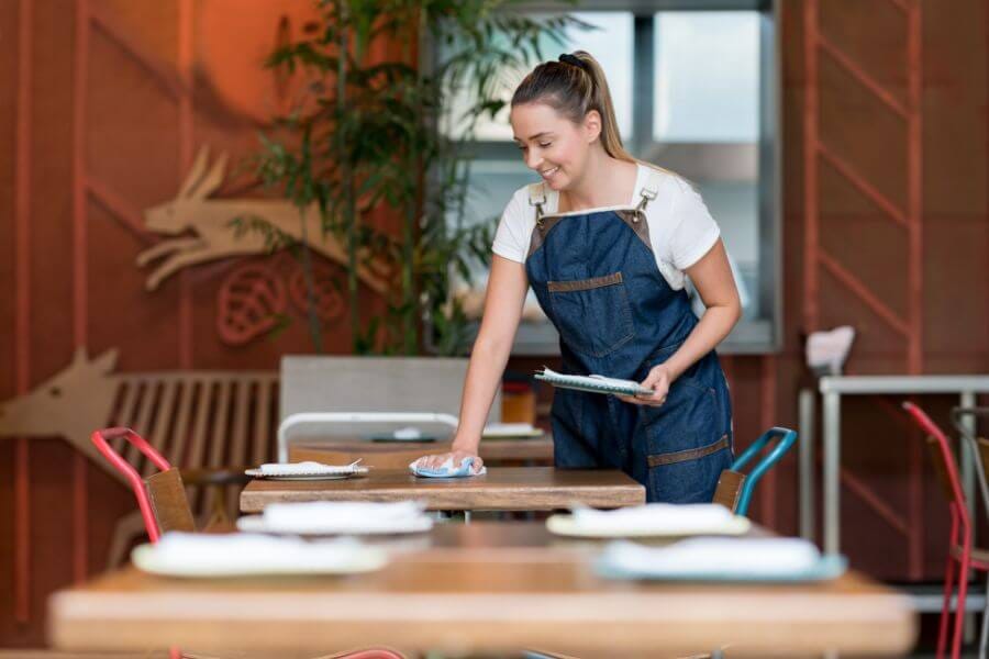 Restaurant server wiping a solid wood butcher-block table top, illustrating the durability, easy sanitation, and guest-appeal benefits of quality commercial restaurant table tops.