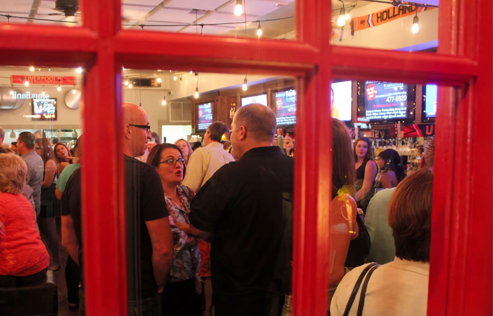 Lively bar crowd at Darby Road Public House in Scotch Plains New Jersey viewed through a red phone booth window frame with Edison bulb string lights and multiple TVs