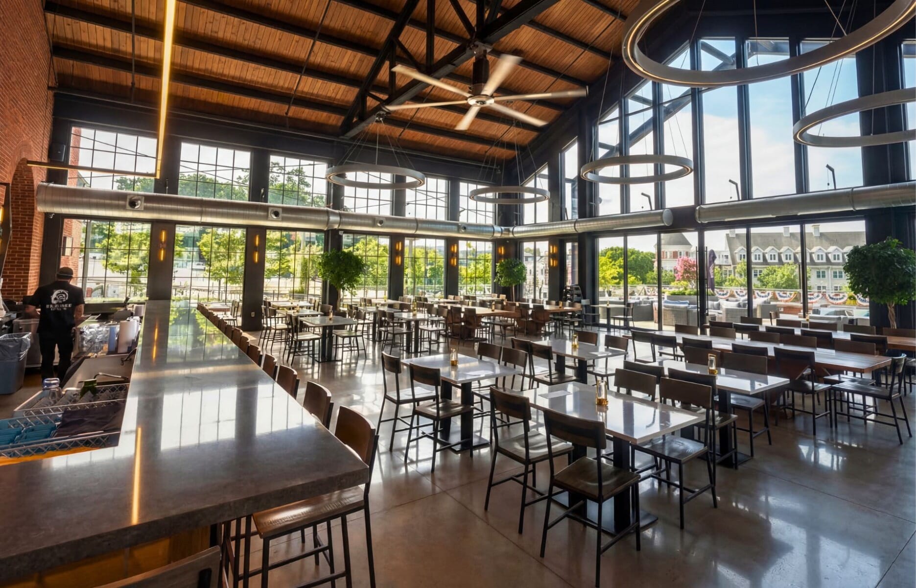 Sunlit dining room at De Cicco and Sons in Sleepy Hollow New York featuring soaring exposed wood beam ceiling, floor-to-ceiling industrial windows, concrete bar, and ring pendant lighting