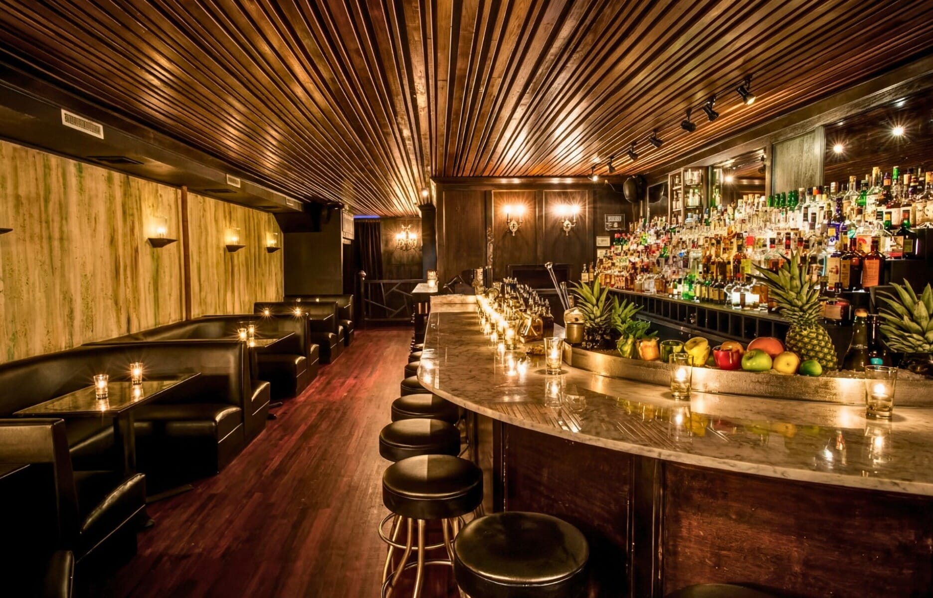 Dimly lit interior of Death and Co cocktail bar in New York City featuring curved marble bar, black leather stools, tufted booths, slatted wood ceiling, and fully stocked back bar