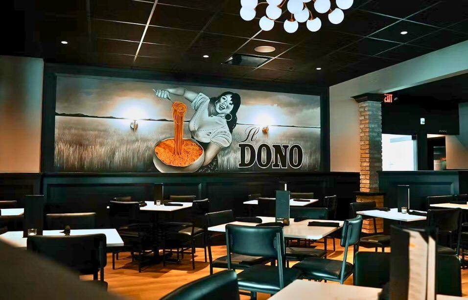 Dining room interior at Il Dono restaurant in Hamilton Township New Jersey featuring large-scale branded pasta mural, black leather chairs, white marble tables, and globe pendant chandelier