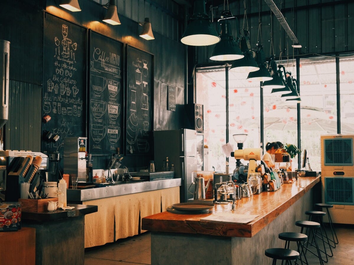 Industrial bar area with backless black metal stools, live-edge wood counter, chalkboard menu walls and factory pendant lights — commercial bar seating in an industrial restaurant