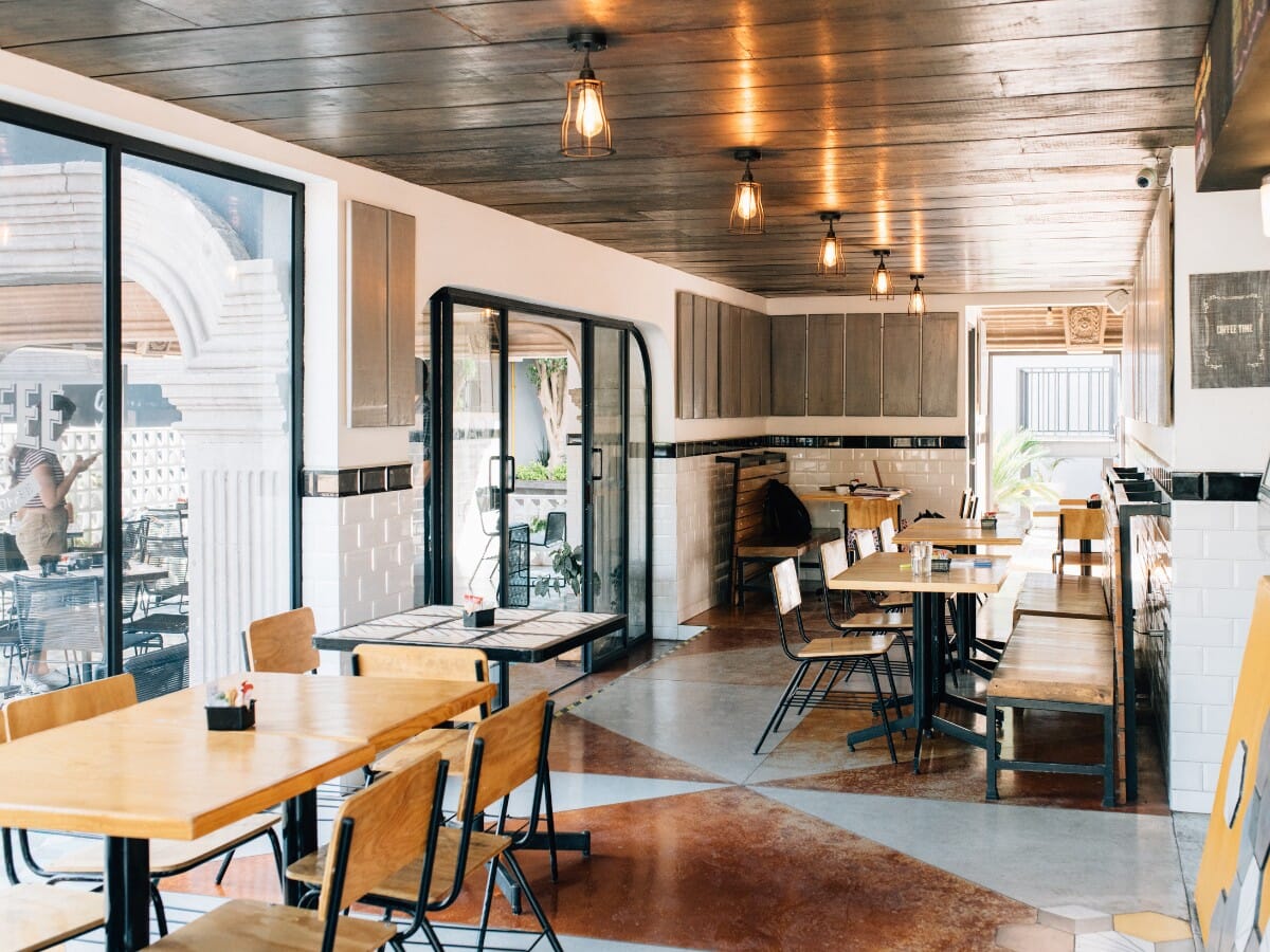Industrial restaurant dining room with matte black steel chairs, solid wood tables, cage Edison pendant lights and white tile walls — commercial industrial furniture in a café setting