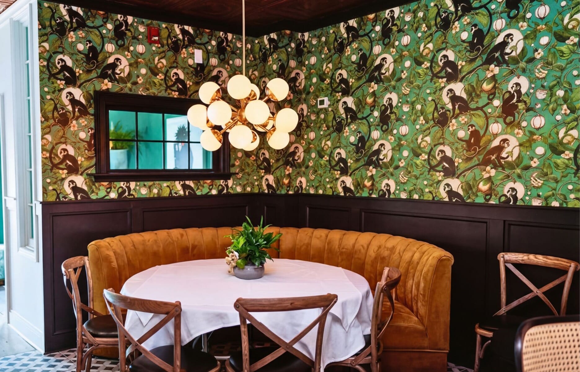 Eclectic dining nook at La Plage Bistro in Bellport New York featuring curved cognac velvet banquette, monkey-print wallpaper, geometric tile floor, and gold globe chandelier