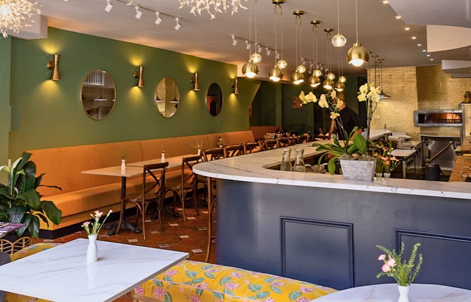 Vibrant dining room interior at Meama restaurant in New York featuring forest green walls with oval gold mirrors, curved navy marble bar, cognac leather banquette, and gold pendant lighting