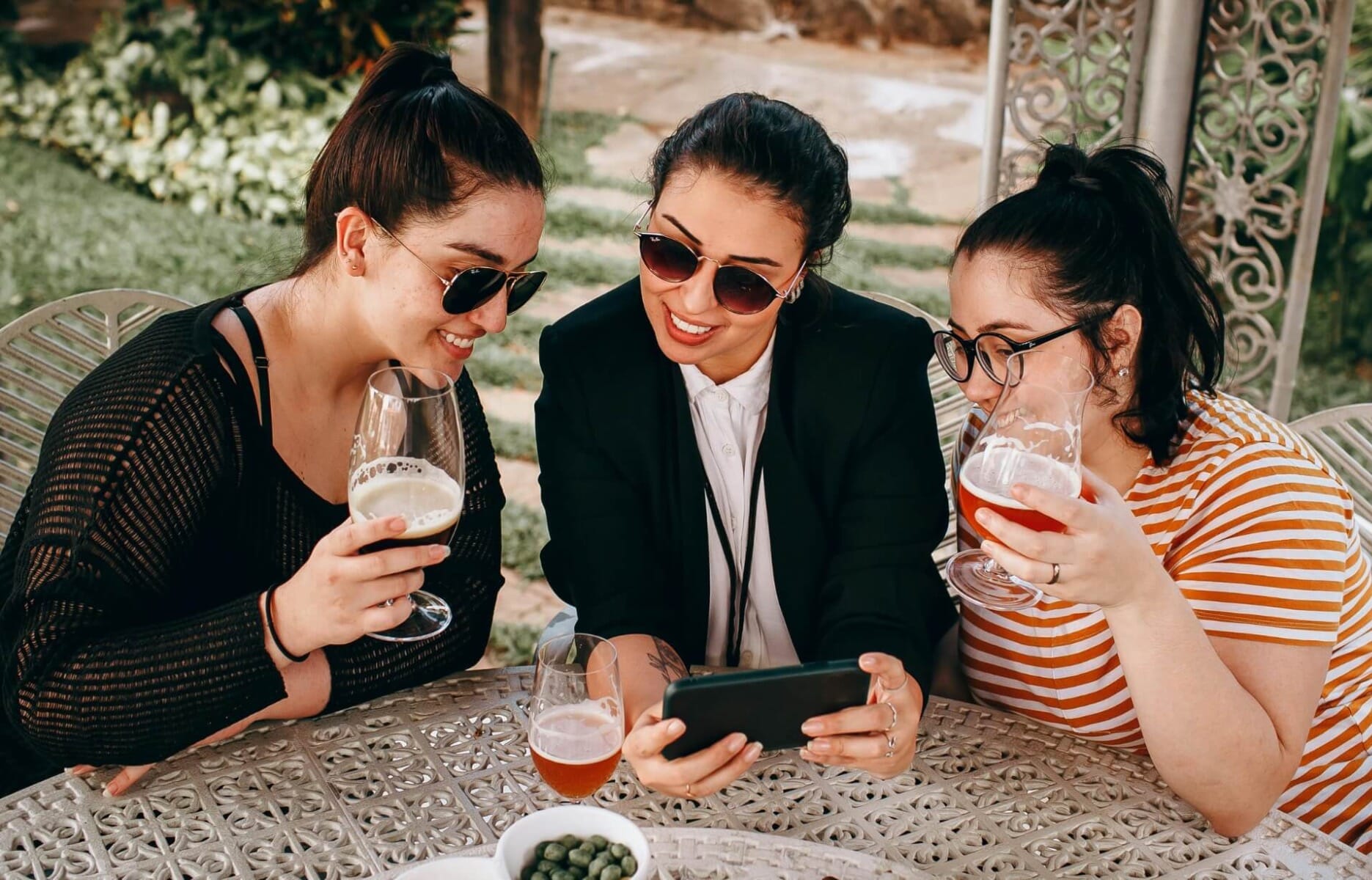Three friends at an outdoor restaurant patio looking at a smartphone together while enjoying drinks — social media discovery is now how most diners find new restaurants