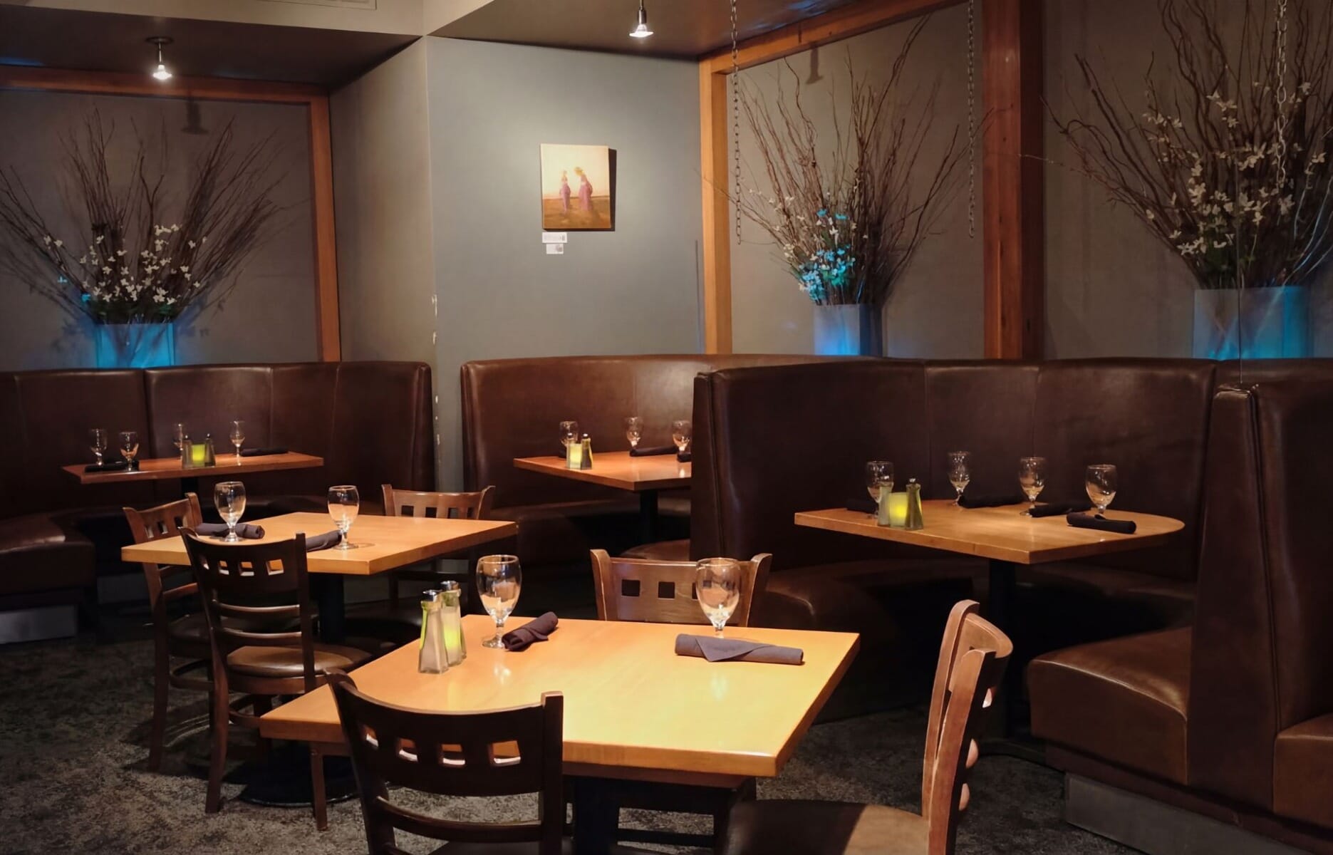 Cozy dining room interior at The Cellar restaurant and wine bar in Corning New York featuring dark brown leather booths, warm wood tables, and blue ambient uplighting with branch arrangements