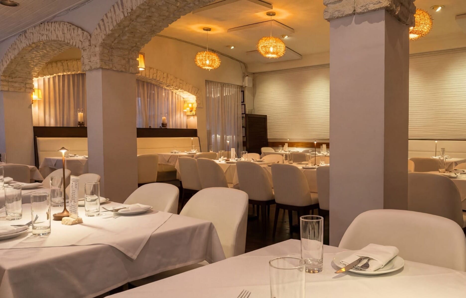 Elegant dining room at The Greek on Main in Metuchen New Jersey featuring white linen tables, white upholstered chairs, stone archways, and warm rattan pendant lights