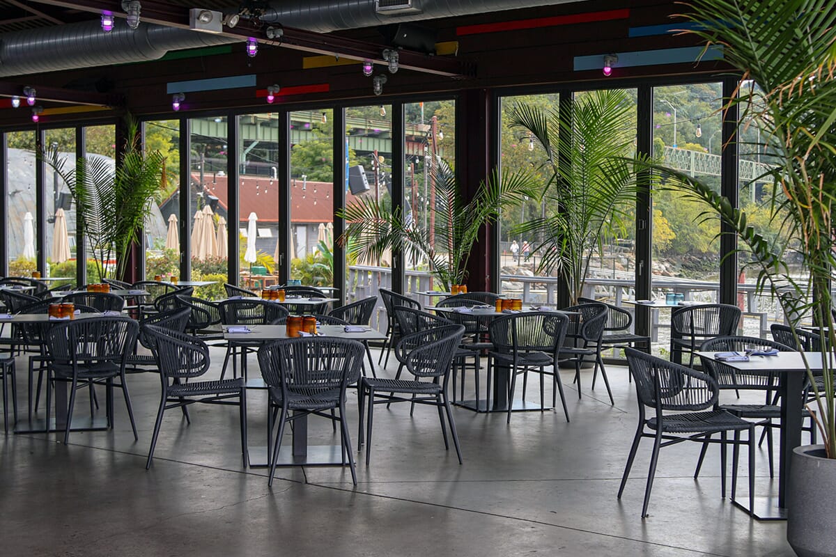 A restaurant with tables and chairs by a large window, offering stunning views of the Hudson River and a vibrant dining experience.