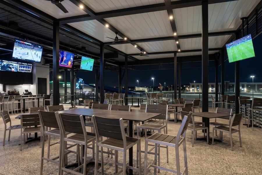 PopStroke Katy outdoor dining area with brushed silver aluminum commercial restaurant tables and pewter synthetic teak seating beside the golf entertainment venue.