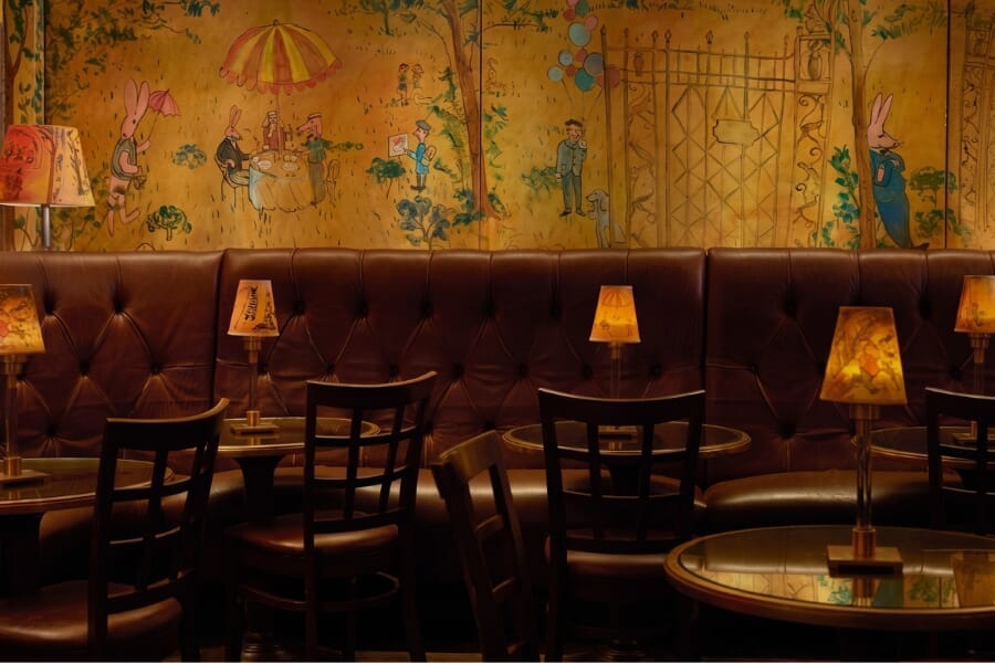 Bemelmans Bar interior at The Carlyle Hotel showing Tea Rose upholstered dark mahogany lattice-back seating, brass details, murals, and an intimate luxury hotel bar atmosphere.