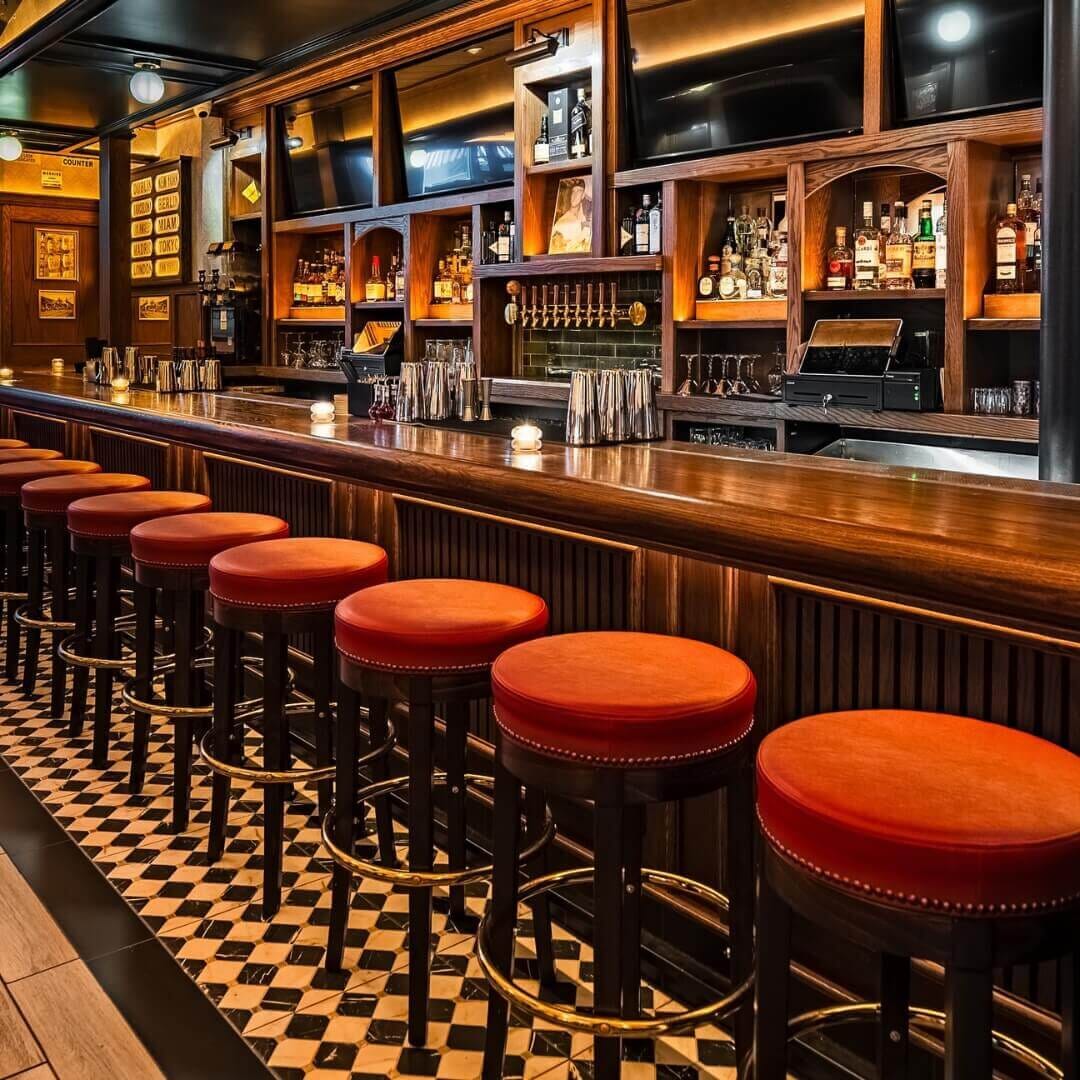 Bar at Central Park Tavern in Midtown NYC featuring walnut counter, red-upholstered bar stools with brass footrests, and classic checkered flooring.
