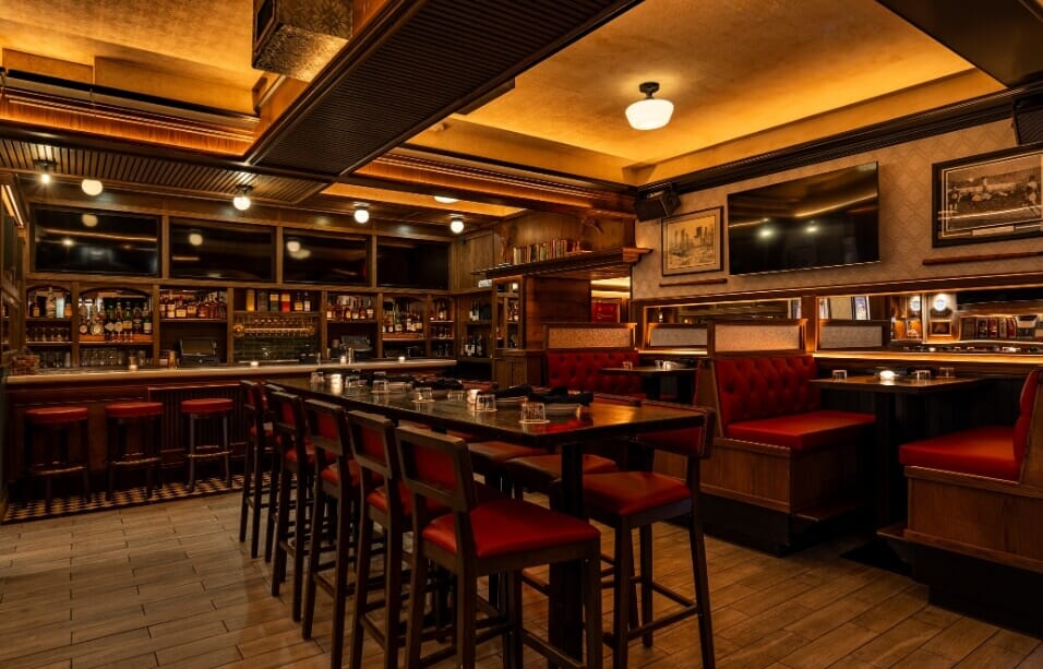 Full view of Central Park Tavern in Midtown NYC showcasing walnut bar rail, brass-accented bar stools, and high-top dining tables under a gold coffered ceiling.
