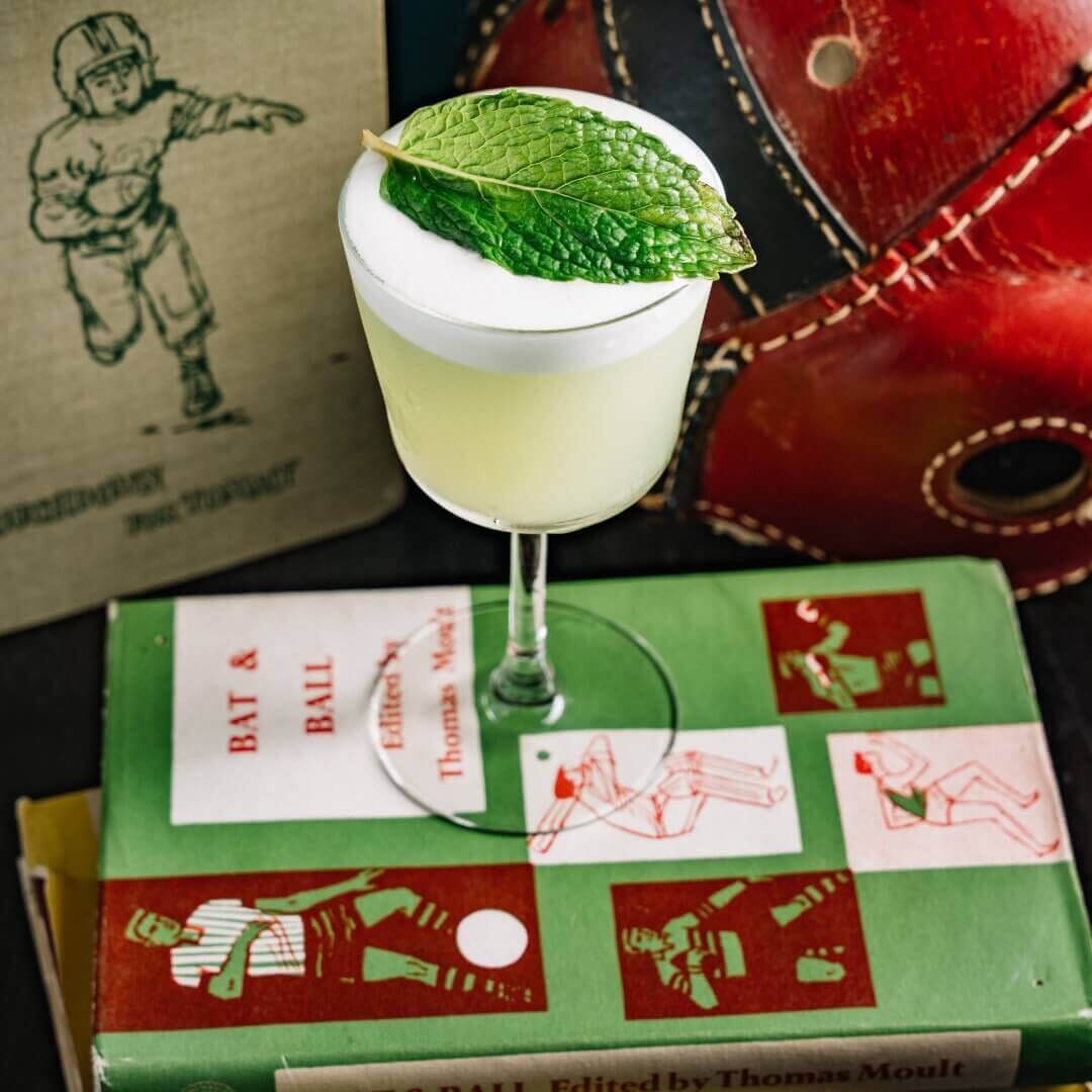 Central Park Tavern NYC signature mint-garnished cocktail photographed beside vintage sports books and red leather restaurant booth seating.