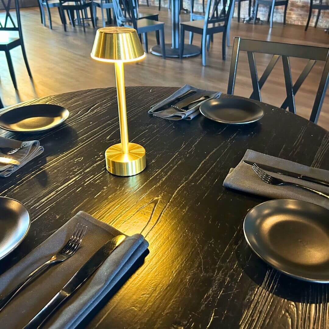 Black oak round table with gold cordless lamp, black plates and X-back chairs at Strickland’s Steakhouse.