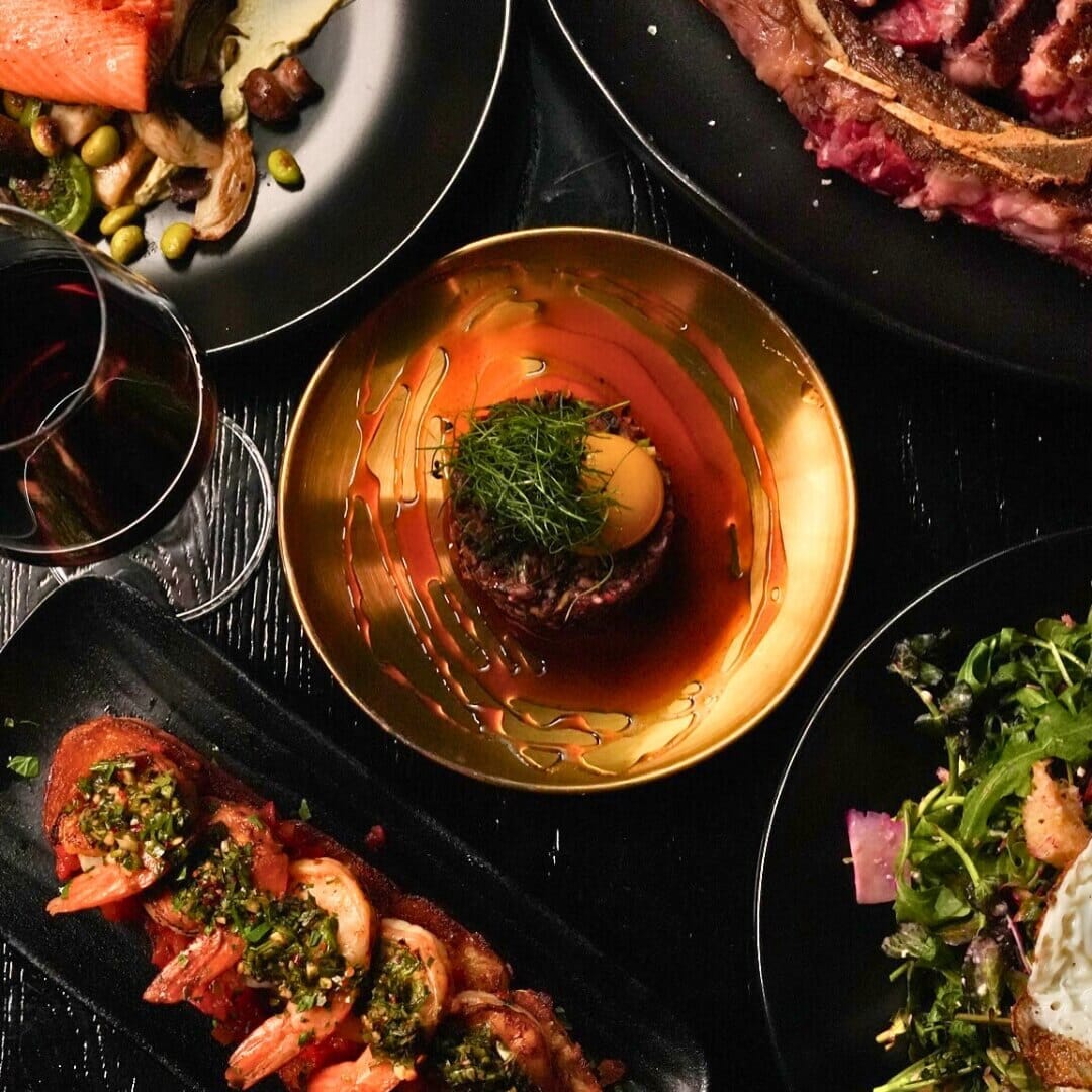 Overhead spread of steak tartare, shrimp toast, rib-eye and salad on black oak table at Strickland’s Steakhouse.