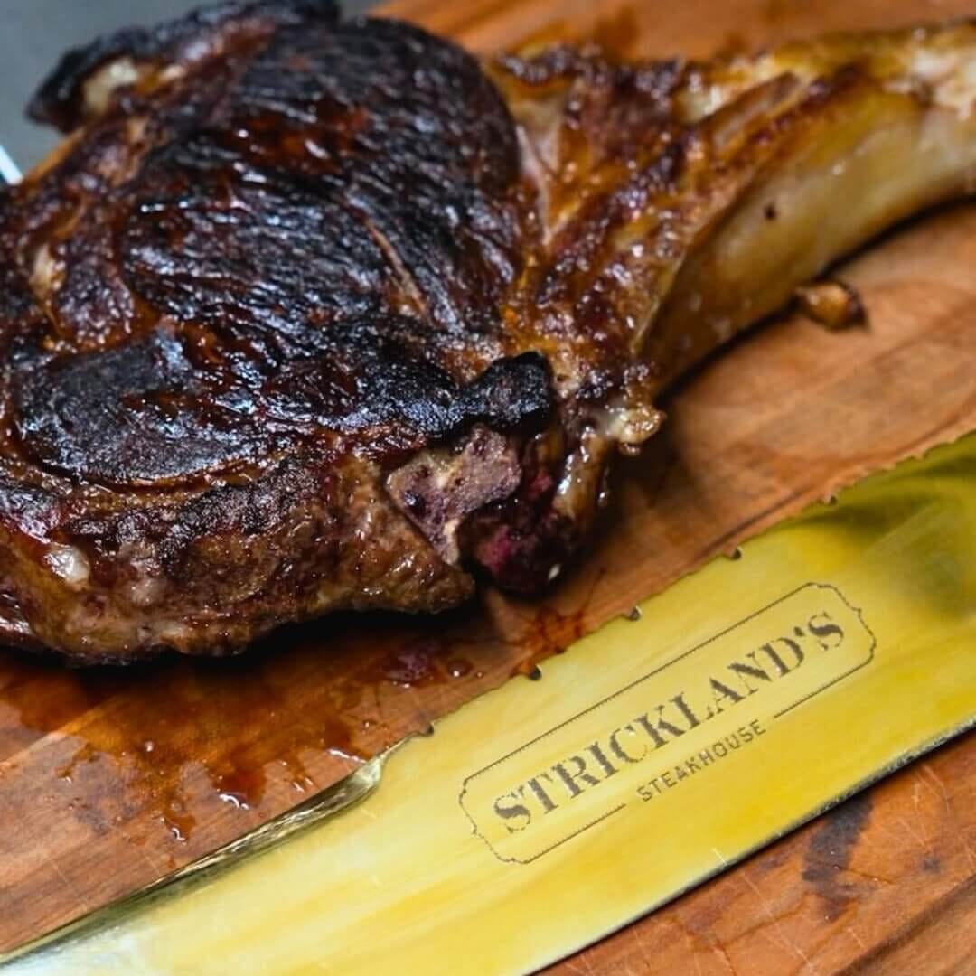 Charred tomahawk rib-eye resting on board with gold Strickland’s Steakhouse knife.
