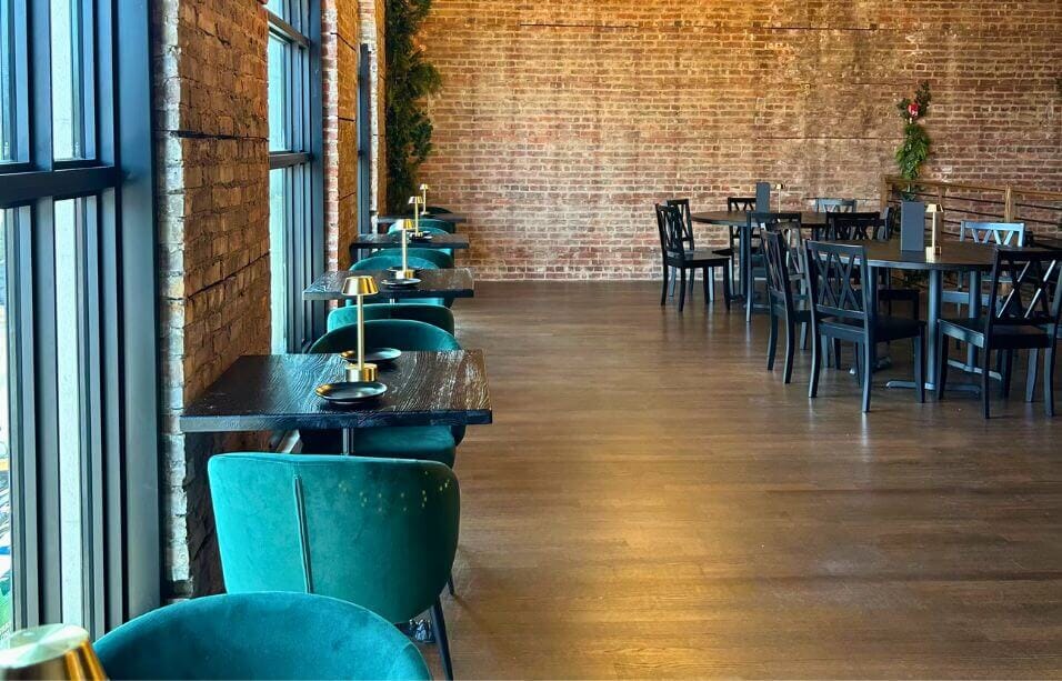 Row of two-top black oak tables with gold lamps, green velvet chairs and exposed-brick windows at Strickland’s Steakhouse.