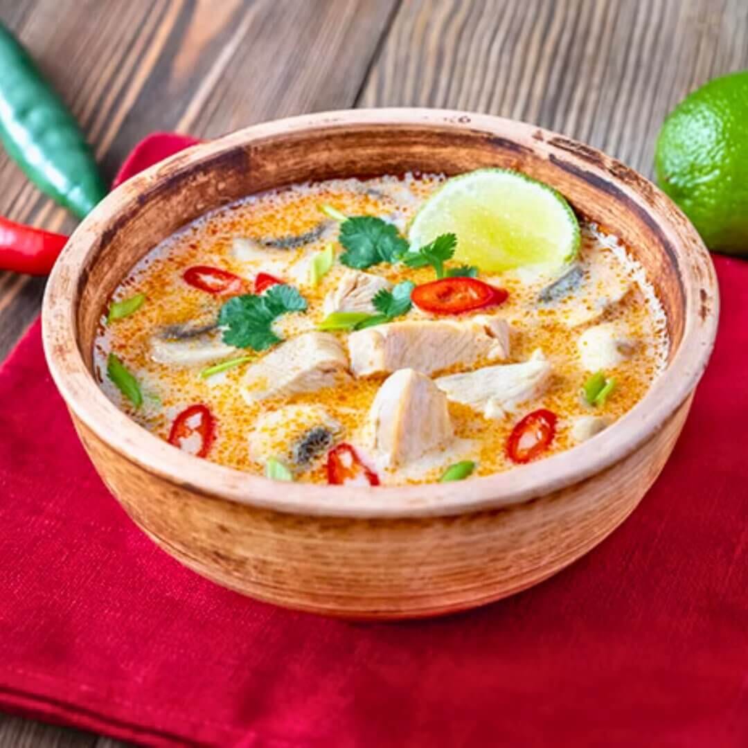 Tom kha coconut chicken soup in wooden bowl with lime wedge, cilantro and red chili slices.