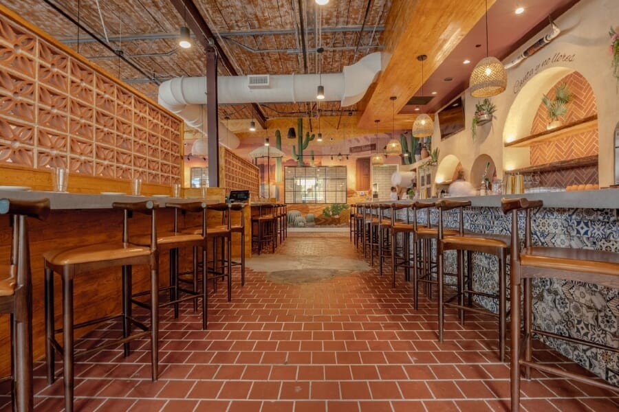 De Nada Birmingham dining room with antique walnut restaurant chairs, tan upholstered seats, colorful desert murals, terracotta tile floor, large windows, and woven pendant lights.
