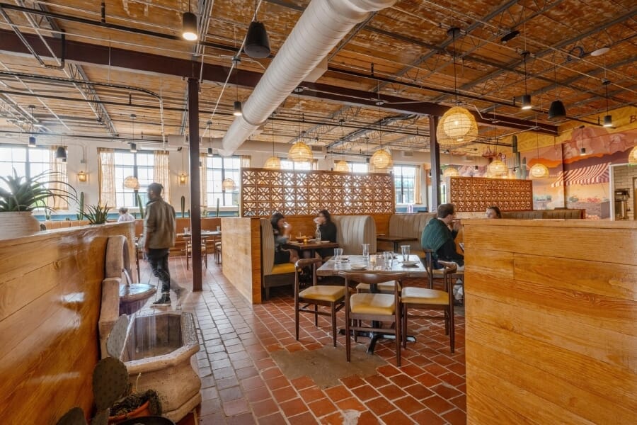 Bright De Nada Birmingham restaurant interior showing warm walnut seating, colorful Mexican-inspired murals, striped booths, terracotta tile, exposed industrial ceiling, and natural woven lighting.