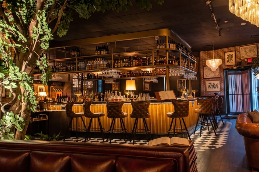Brown tufted bucket bar stools arranged along a warmly lit restaurant bar with brass accents, greenery, and checkered flooring.