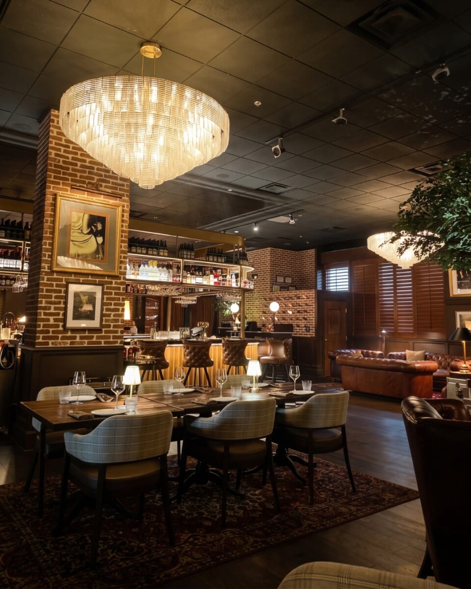 The Belmont modern American steakhouse interior with burgundy leather booths, patterned armchairs, reclaimed wood tables, brick columns, and dramatic chandeliers.