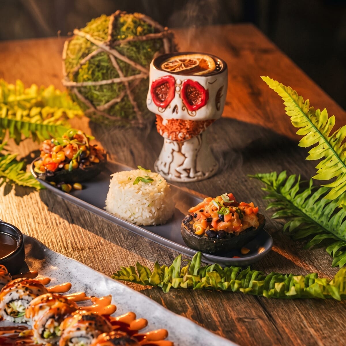 Sushi rolls, filled avocado halves, rice, and a skull-shaped cocktail vessel on a wood table at Vida.